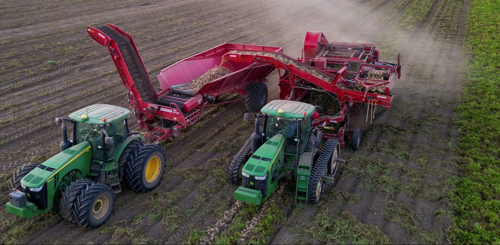 Dois carrinhos de colheita e dois tratores John Deere sendo usados em uma fazenda para carregar batatas colhidas.
