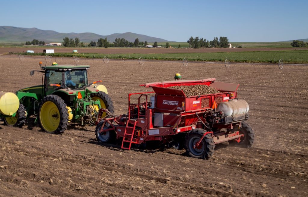 A plantadeira de batatas 8046 - 6 Row Potato Bed da Spudnik e um trator John Deere sendo usado em uma fazenda.