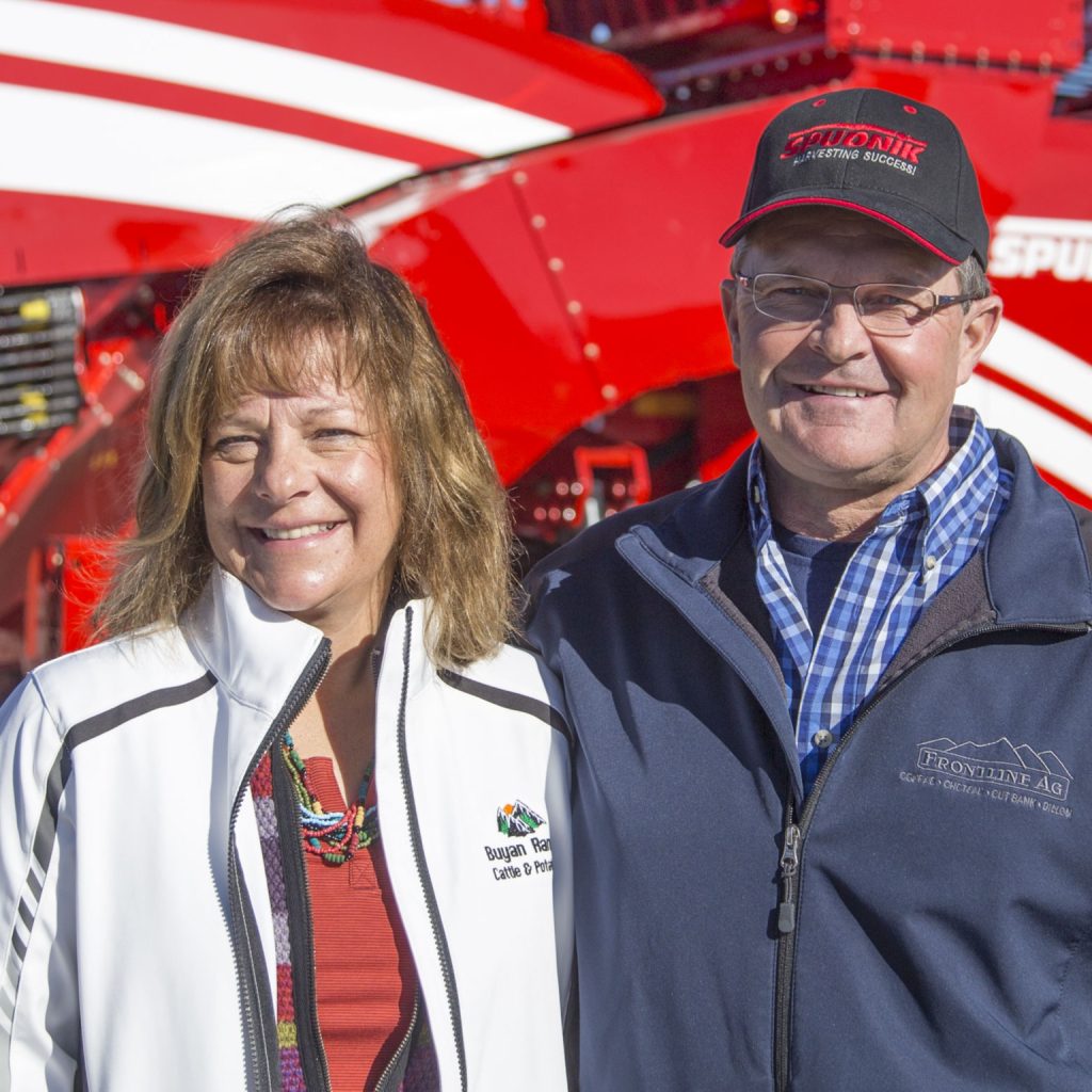 A couple: a man and a woman, both white people, standing in front of a Spudnik piece of farming equipment.